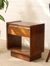 Load image into Gallery viewer, A mid-century modern wooden nightstand with a single drawer, open shelf, and block base, shown in a warm brown finish against a white brick wall.
