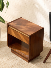 Load image into Gallery viewer, A mid-century modern wooden nightstand with a warm brown finish, featuring a single drawer with a light wood trim and an open shelf below, set on a textured beige rug against a white brick wall.
