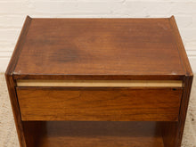 Load image into Gallery viewer, A mid-century modern wooden nightstand with a warm brown finish, featuring a drawer with a light wood accent strip and an open shelf below, set against a white brick wall.
