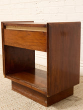 Load image into Gallery viewer, A mid-century modern wooden nightstand with a single drawer and open shelf, shown against a white brick wall and light carpet.
