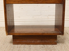 Load image into Gallery viewer, A close-up of a dark brown wooden furniture piece, showing a lower shelf and block-style base against a white brick wall and beige woven rug.
