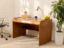 Load image into Gallery viewer, A mid-century modern wooden writing desk with a slanted top, set in a bright room against a white brick wall. The desk is styled with a silver desk lamp, a yellow rotary phone, books, and a small clock, with a potted plant and office chair nearby.

