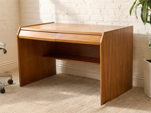 Load image into Gallery viewer, A mid-century modern wooden desk with a warm brown finish, featuring a top drawer, open shelf, and angled legs, set against a white brick wall and beige carpet.
