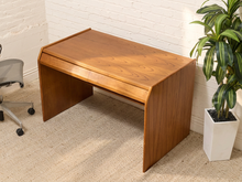 Load image into Gallery viewer, A mid-century modern wooden desk with a warm walnut finish, positioned in a room corner against a white brick wall with a potted plant and office chair visible.
