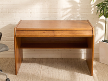 Load image into Gallery viewer, A mid-century modern wooden writing desk with a warm medium-brown finish, featuring a single drawer, an open shelf, and angled legs, set against a white brick wall.

