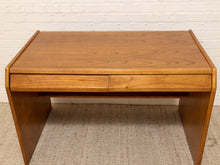 Load image into Gallery viewer, A mid-century modern wooden desk with a warm medium-brown finish, featuring a top drawer and angled side supports, set against a white brick wall and textured beige rug.

