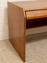 Load image into Gallery viewer, A close-up of a mid-century modern wooden desk or console table with a warm medium-brown finish, featuring a drawer and open shelf, set against a white brick wall and beige carpet.
