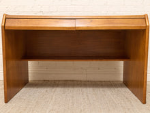 Load image into Gallery viewer, A mid-century modern wooden desk with a warm medium-brown finish, featuring a top drawer, an open shelf, and angled side supports, set against a white brick wall and light beige carpet.
