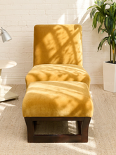 Load image into Gallery viewer, A mustard yellow velvet lounge chair with a matching ottoman and dark wood base, set against a white brick wall with dappled sunlight and a potted plant in the background.
