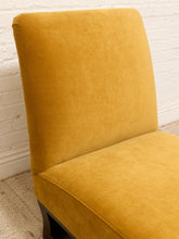 Load image into Gallery viewer, A close-up of a mustard yellow velvet upholstered chair with a high back, positioned against a white brick wall and light beige rug.
