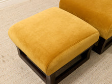 Load image into Gallery viewer, A mustard yellow velvet ottoman with a dark wood base, shown alongside another matching ottoman on a textured beige rug.
