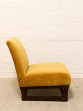Load image into Gallery viewer, A mustard yellow velvet accent chair with a dark wood frame, positioned against a white brick wall and light beige carpet.
