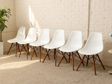 Load image into Gallery viewer, Six modern white molded plastic chairs with tapered brown wooden legs arranged in a row against a white brick wall on a beige carpet.
