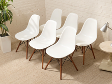 Load image into Gallery viewer, Six white molded plastic side chairs with tapered brown wooden legs and black metal X-base supports, arranged on a beige carpeted floor against a white brick wall.
