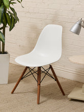 Load image into Gallery viewer, A modern white molded plastic side chair with tapered wooden legs and black metal cross-members, shown in a styled room setting with a brick wall, potted plant, and lamp.
