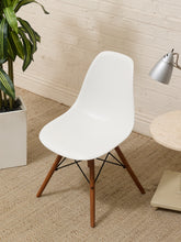 Load image into Gallery viewer, A modern white molded plastic side chair with tapered wooden legs and black metal cross-members, shown in a styled room setting with a brick wall, carpet, a potted plant, and a side table.
