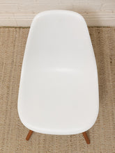 Load image into Gallery viewer, A white molded plastic chair with tapered wooden legs, photographed from a top-down angle on a woven jute rug against a white brick wall.
