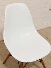 Load image into Gallery viewer, A modern white molded plastic chair with tapered wooden legs, shown against a white brick wall and beige rug.
