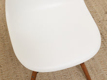 Load image into Gallery viewer, Close-up of a white molded plastic chair seat with light brown wooden legs on a beige textured rug.
