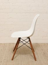 Load image into Gallery viewer, A modern side chair with a white molded plastic shell and tapered wooden legs, shown against a white brick wall and light beige rug.
