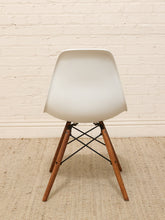 Load image into Gallery viewer, A modern molded plastic side chair with a white seat and wooden legs, positioned against a white brick wall on a textured beige rug.

