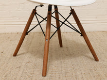 Load image into Gallery viewer, A close-up of a mid-century modern chair showing its white molded plastic seat, brown wooden legs, and black metal cross-struts, set on a woven beige rug against a white brick wall.
