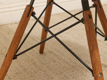 Load image into Gallery viewer, Close-up of a chair leg assembly with medium brown wooden legs and black metal cross-bracing on a beige carpeted floor.
