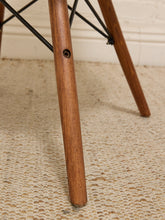 Load image into Gallery viewer, Close-up of a mid-century modern wooden chair leg with a medium brown wood finish and a black metal Eames-style DSW base, resting on a textured beige woven rug.
