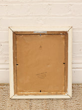 Load image into Gallery viewer, The back of a vintage framed picture showing a distressed off-white wooden frame, brown cardboard backing, and a small metal hanger at the top, set against a white brick wall and woven rug.
