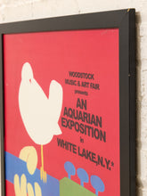 Load image into Gallery viewer, A framed poster print featuring a bold red background with a large white dove, colorful abstract shapes, and text reading 'Woodstock Music &amp; Art Fair presents An Aquarian Exposition in White Lake, N.Y.
