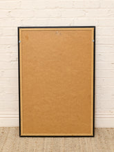 Load image into Gallery viewer, A large rectangular cork board with a black frame and a tan inner border, leaning against a white painted brick wall on a woven rug.
