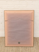 Load image into Gallery viewer, The back of a vintage rectangular framed picture with a peach-colored backing and a metal wire hanger, leaning against a white brick wall on a woven rug.
