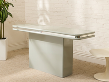 Load image into Gallery viewer, A modern mid-century style dining table with a glossy pale green top and a matte sage green pedestal base, set against a white brick wall and beige carpet.
