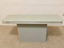 Load image into Gallery viewer, A mid-century modern low coffee table with a light sage green painted metal frame and a rectangular pedestal base, set on a woven jute rug against a white brick wall.
