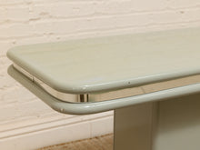 Load image into Gallery viewer, Close-up of a glossy pale sage green table corner with a chrome metal trim strip, set against a white brick wall background.
