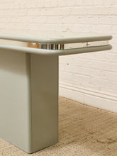 Load image into Gallery viewer, A close-up of a mid-century modern table leg and corner edge, featuring a sage green painted finish, chrome metal trim, and a clear glass top, set against a white brick wall and beige rug.
