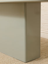 Load image into Gallery viewer, Close-up of a light sage green metal desk leg or cabinet corner standing on a beige woven rug against a white brick wall.
