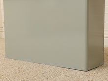Load image into Gallery viewer, Close-up of the corner of a muted sage green metal cabinet or utility box standing on a beige carpeted floor against a white brick wall.

