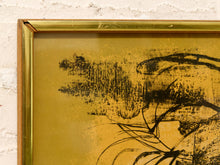 Load image into Gallery viewer, Close-up of a framed abstract art print featuring black ink brushstrokes on a textured gold background, set in a gold-toned frame against a white textured wall.
