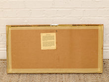 Load image into Gallery viewer, The back of a framed artwork or print, showing a tan cardboard backing with a small paper label attached, set against a white painted brick wall and woven rug.
