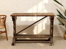 Load image into Gallery viewer, A dark brown wooden console table with ornate turned legs, a diagonal support brace, and casters, set against a white brick wall.
