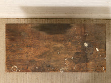 Load image into Gallery viewer, A rectangular distressed wooden board with a dark brown finish, showing signs of wear, scratches, and peeling paint, placed on a beige woven rug against a white brick wall.
