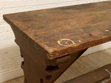 Load image into Gallery viewer, A close-up of a rustic dark brown wooden table or bench with a thick top and carved trestle-style legs, set against a white brick wall and light carpet.
