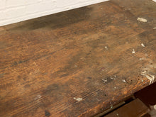Load image into Gallery viewer, A close-up of a worn, dark brown wooden tabletop with visible grain, scratches, chips, and peeling paint, set against a white painted brick wall.
