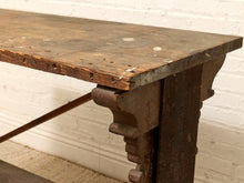 Load image into Gallery viewer, Close-up of a rustic dark wood and metal table corner with decorative bracket supports, set against a white painted brick wall.
