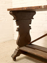 Load image into Gallery viewer, A close-up of a dark brown wooden table leg with carved details and a rustic wood tabletop, set against a white brick wall and light carpet.
