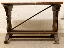 Load image into Gallery viewer, An antique dark wood console table with ornate turned legs, a diagonal support brace, and caster wheels, set against a white brick wall.
