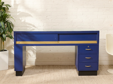 Load image into Gallery viewer, A modern blue writing desk with a gold horizontal accent stripe, featuring a two-drawer top section, a three-drawer pedestal on the right, and black legs, set against a white brick wall.
