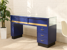 Load image into Gallery viewer, A mid-century modern writing desk in royal blue with a gold horizontal accent stripe, a glass top, three drawers on the right, and black legs, shown in a styled room setting with a white brick wall, beige carpet, a potted plant, and a chair.
