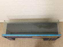 Load image into Gallery viewer, A rectangular glass display case or aquarium with a dark blue wooden base, positioned on a beige woven rug against a white brick wall.

